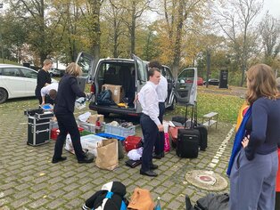 Abbildung einer Gruppe von Personen beim Sortieren von Gegenständen rund um offenen Transporter auf Parkplatz.