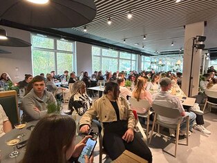Abbildung von einem vollen Saal mit Menschen an Tischen sitzen.