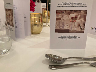 Abbildung einer festlich gedeckten Tafel mit Besteck, Wasserglas und Tischkarte mit Infos zum Wettbewerbsessen 2024.