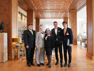 Abbildung einer Gruppe von sechs Personen in festlicher Kleidung in elegantem Innenraum.
