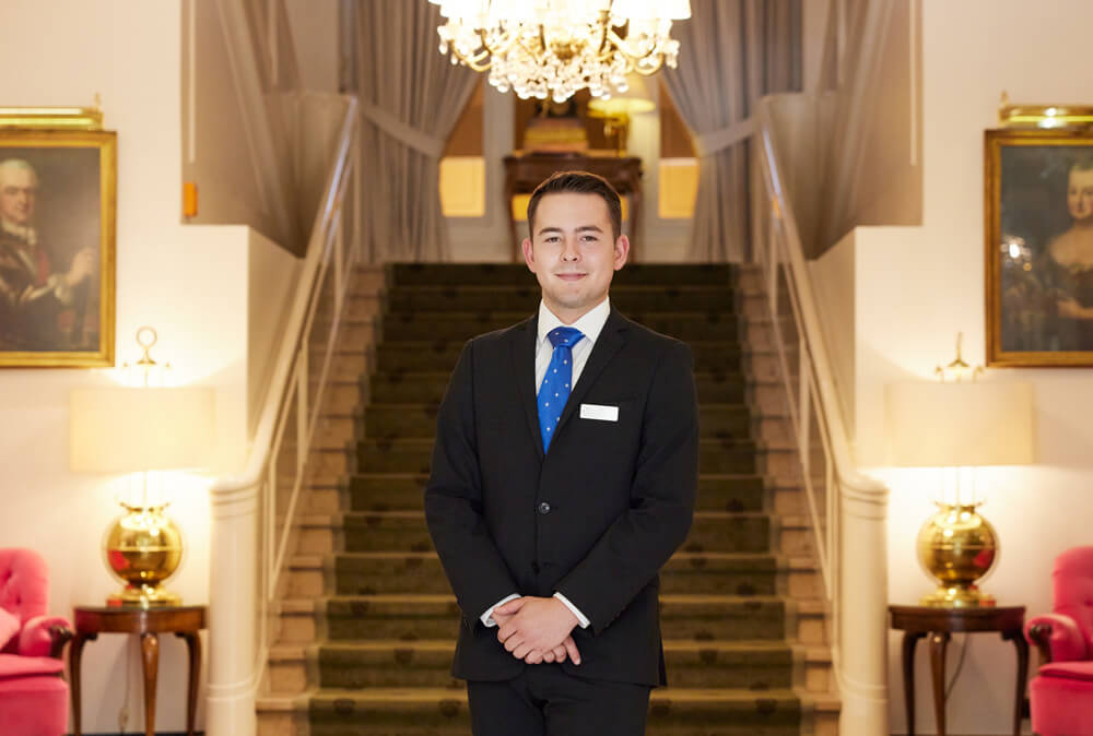 Abbildung eines Mannes im Anzug in einer Hotellobby vor einer großen Treppe im Hintergrund.
