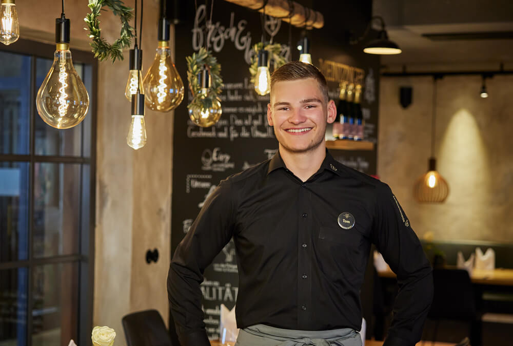 Abbildung eines Kellners in schwarzer Uniform, der in einem Restaurant vor einer Wand mit Schiefertafel steht und in die Kamera lächelt.