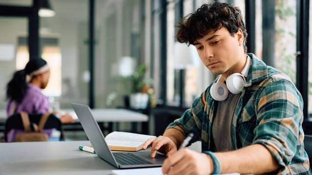 Abbildung eines Jungen mit Laptop, Buch und Kopfhörern beim Schreiben an einem Tisch.