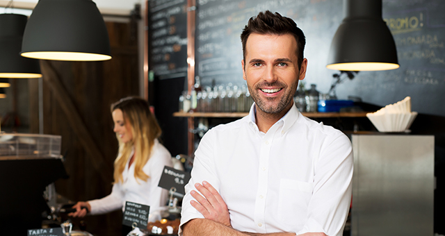 Abbildung von zwei Personen in einem Café, vorne Person mit verschränkten Armen, im Hintergrund Theke mit Flaschen, Gläsern und Kreidetafel.