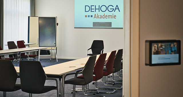 Abbildung eines Konferenzraums mit Tischen und Stühlen, vorne Leinwand.