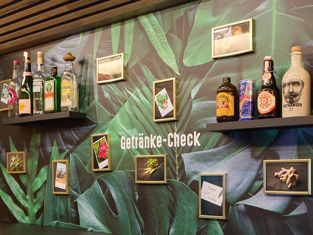 Abbildung einer Wand mit dem Schriftzug „Getränke-Check“, dekoriert mit Bildern und Flaschenregalen.