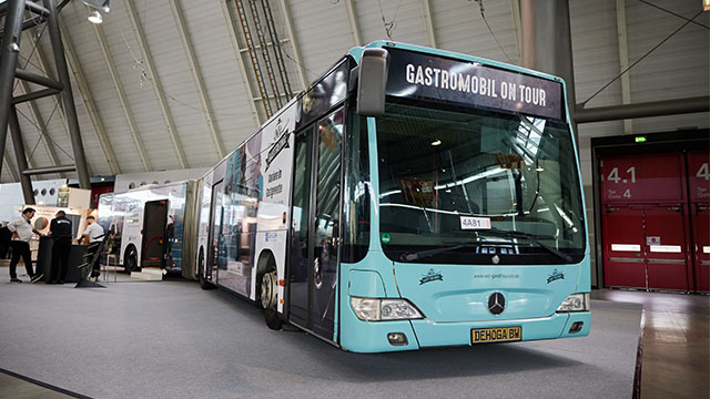 Abbildung eines Busses mit der Aufschrift „GASTROMOBIL ON TOUR“ in einer Ausstellungshalle.