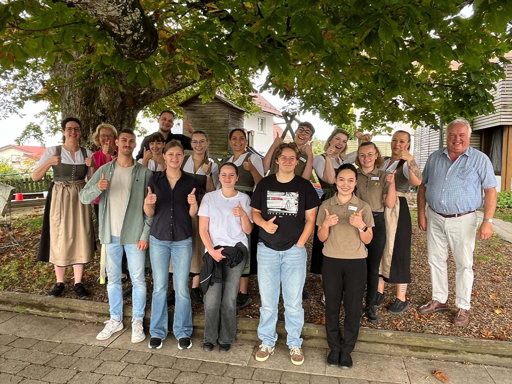 Abbildung von 15 Personen unter einem großen Baum, teils in traditioneller Kleidung, posierend mit Daumen-hoch-Geste.