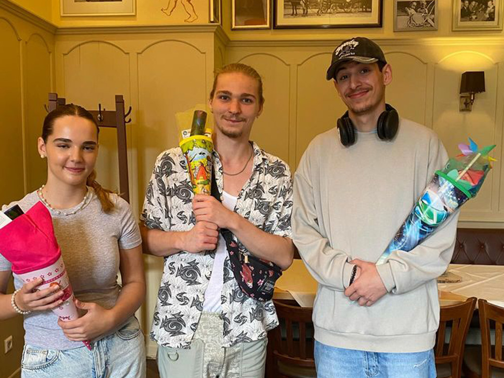 Abbildung von drei Personen mit bunten Schultüten in einem Innenraum mit Bildern an der Wand und Holzmöbeln.