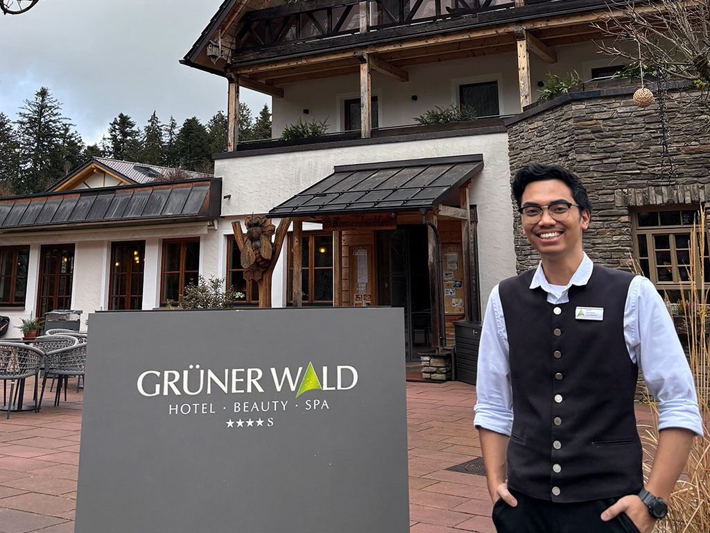 Abbildung eines Hotels mit dem Schild „GRÜNER WALD HOTEL • BEAUTY • SPA *****S“ und einer Person in Uniform daneben.