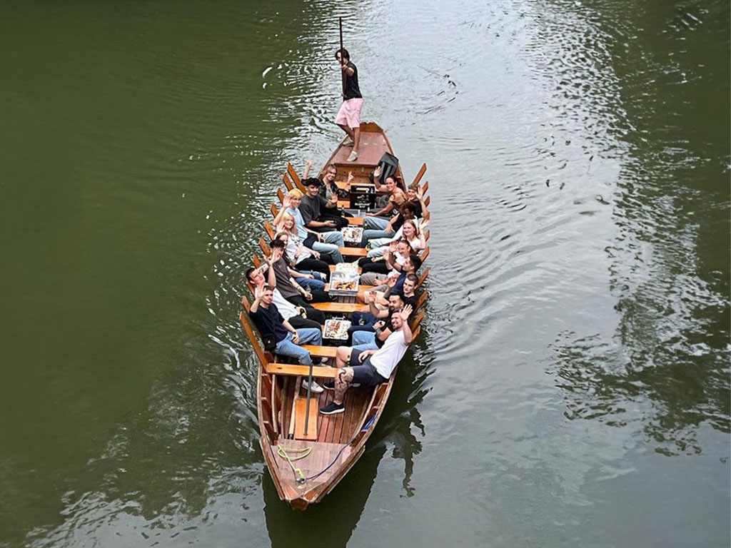Abbildung einer Gruppe auf einem Holzboot mit Speisen und Getränken, gesteuert von einer Person mit Stange auf einem Gewässer.