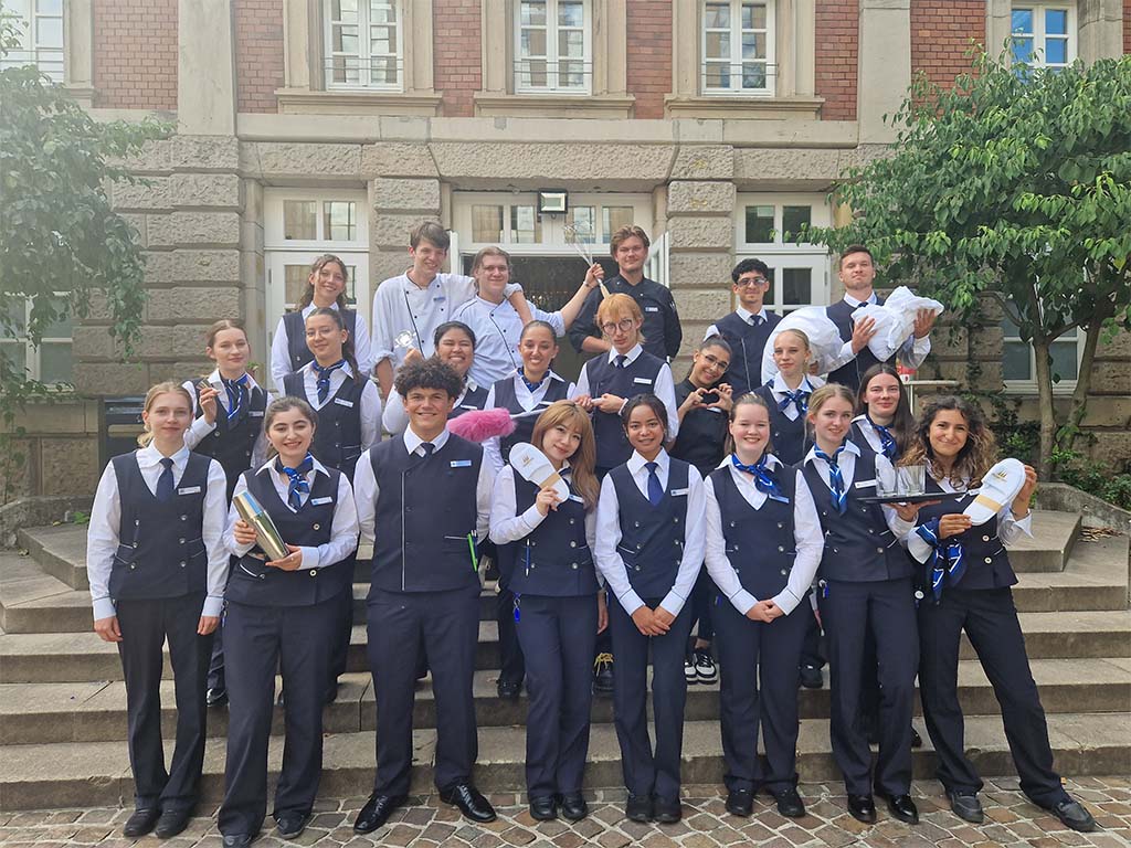 Abbildung einer Gruppe in Uniform und Kochkleidung auf Treppenstufen vor einem Gebäude mit Backsteinfassade und Grünpflanzen.