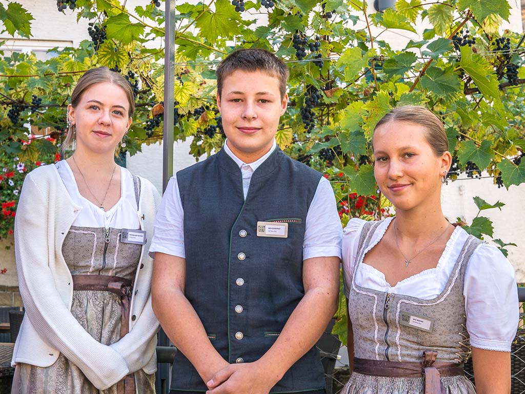 Abbildung von drei Personen in traditioneller Kleidung vor Weinreben mit sichtbaren Trauben.