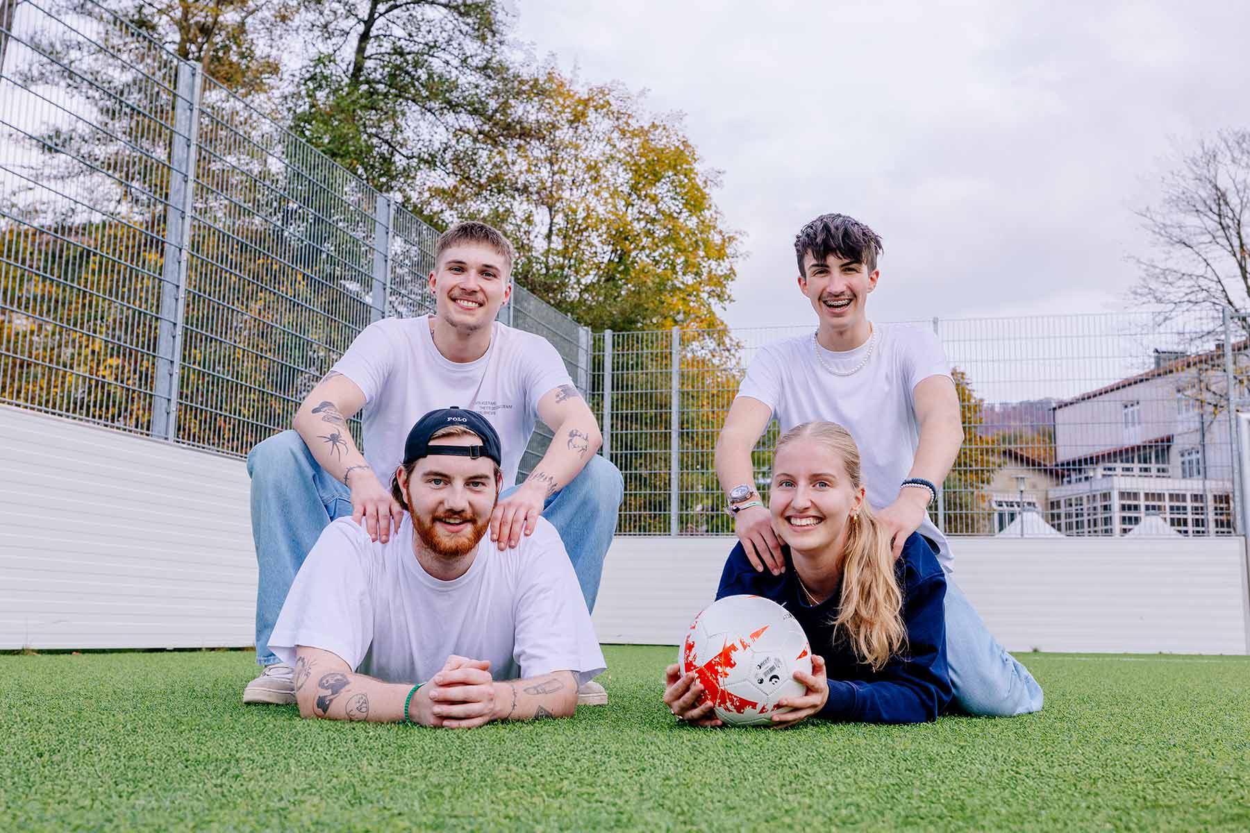 Abbildung von vier Personen auf einem Fußballfeld, zwei liegen, zwei knien dahinter, einer hält einen Ball.