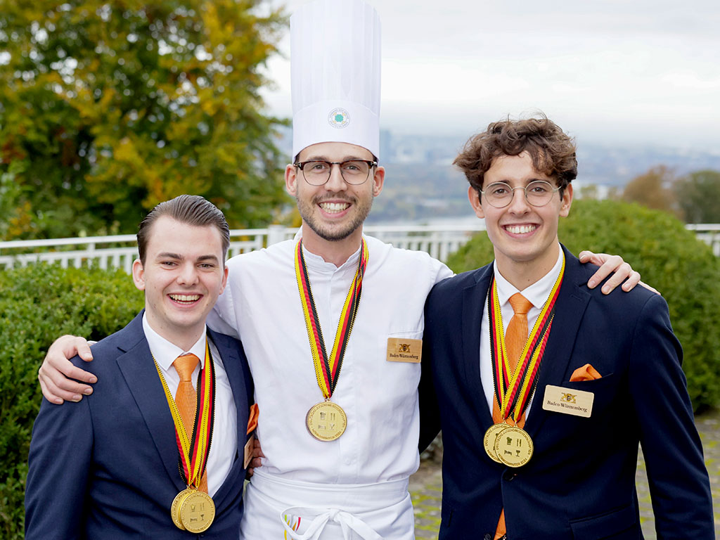 Abbildung von drei Männern im mit Goldmedaillen im Freien, mittig ein Koch in Uniform, flankiert von zwei Personen in Anzügen.