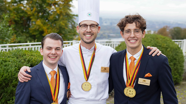Abbildung von drei Männern im mit Goldmedaillen im Freien, mittig ein Koch in Uniform, flankiert von zwei Personen in Anzügen.