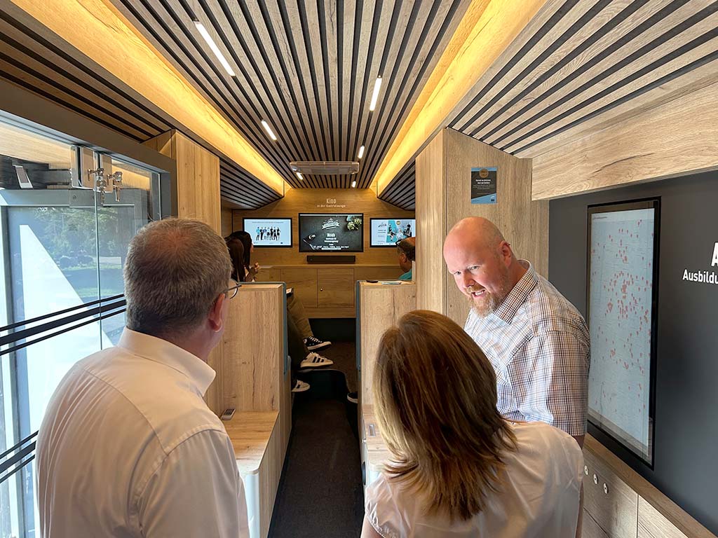 Abbildung von einem modernen mobilen Ausstellungsraum mit Holzverkleidung, Bildschirmen und mehreren Personen. Abbildung von einem modernen mobilen Ausstellungsraum mit Holzverkleidung, Bildschirmen und mehreren Personen.