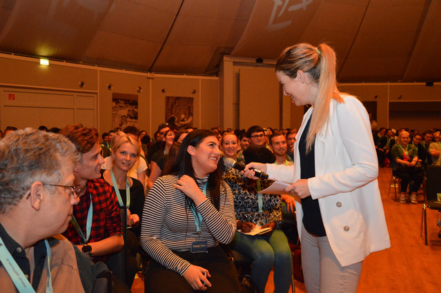 Abbildung von einer Frau mit Mikrofon vor Publikum in einem Konferenzraum.