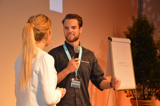 Zwei Personen im Gespräch, eine mit Mikrofon, im Hintergrund ein Flipchart mit Zeichnungen – Szene eines Vortrags oder Workshops.