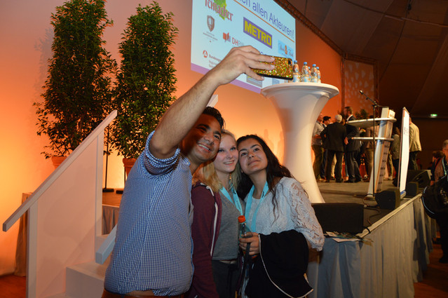 Drei Personen machen ein Selfie vor einer Bühne mit Logos und Pflanzen – Szene eines Events oder einer Konferenz.
