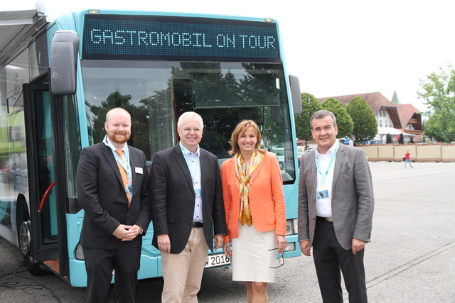 Abbildung vvon vier Personen vor einem Bus im Freien mit Bäumen und Gebäuden.
