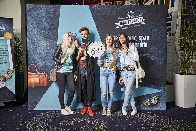Vier Personen vor einem Hintergrund mit „Wir Gastfreunde“ und „Teamwork, Spaß & Zusammenhaltendrin“ – mit Deko wie Taschen, Artischocken und Pflanze.