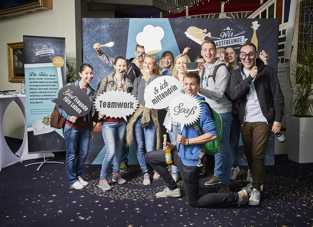 Abbildung einer Gruppe vor Kulisse mit Kochmotiven und Schildern wie „Teamwork“, „Spaß“ und „Echte Freunde fürs Leben“ – mit weiterem Bannertext im Hintergrund.