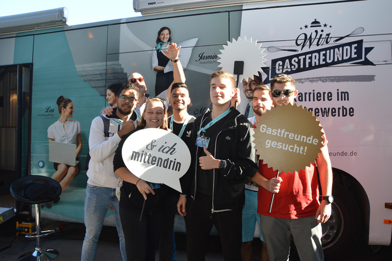 Abbildung einer Gruppe vor einem Promo-Fahrzeug mit Aufschriften wie „Wir GASTFREUNDE“ und „Karriere im Gastgewerbe“.