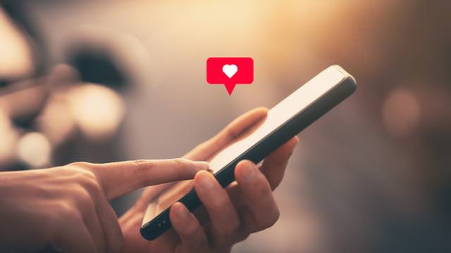 Abbildung einer Hand, die ein Smartphone hält und den Bildschirm berührt, mit einem roten Herzsymbol in einer Sprechblase darüber.